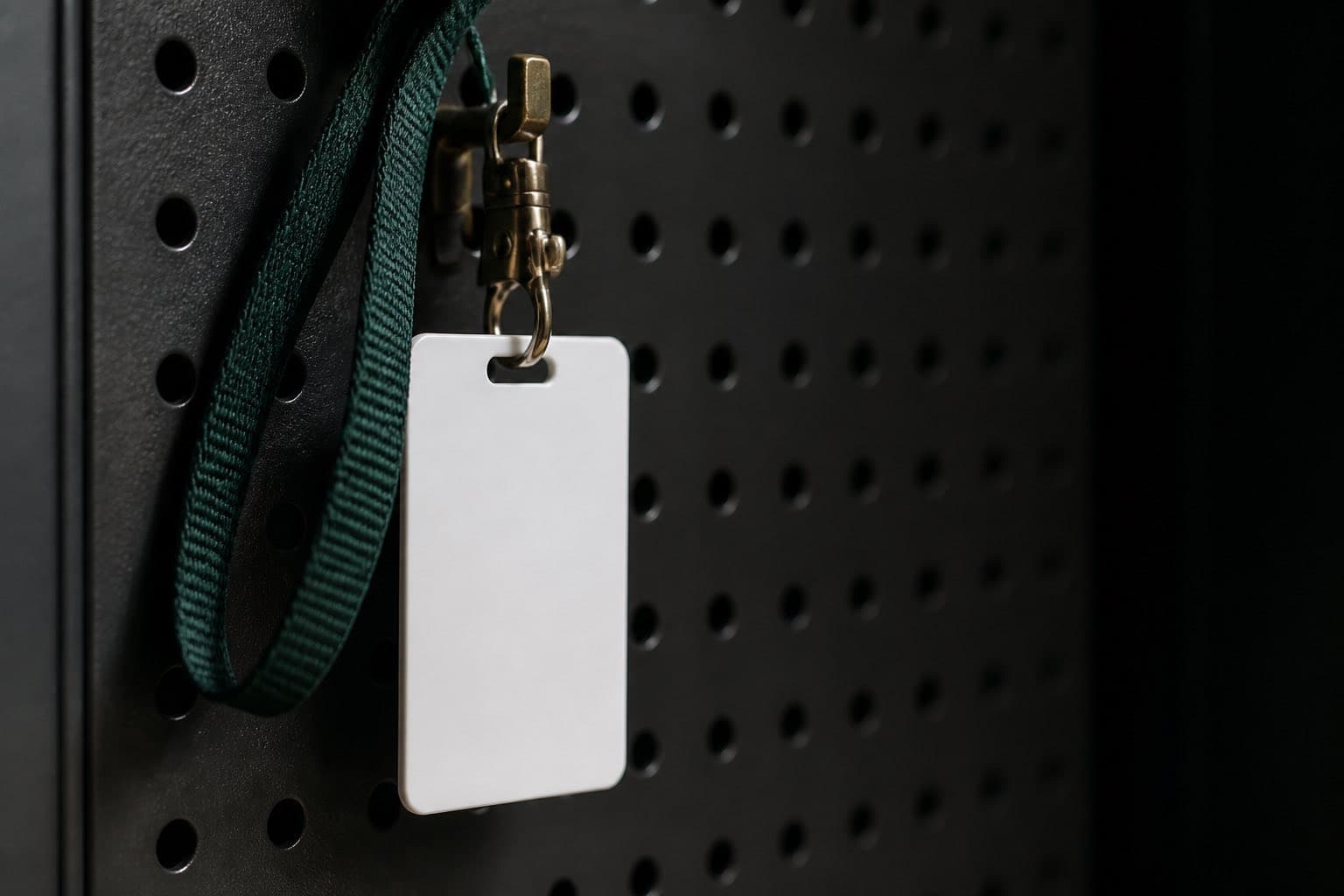 A blank workplace ID badge on a forest-green lanyard hanging from a brass hook on a perforated panel