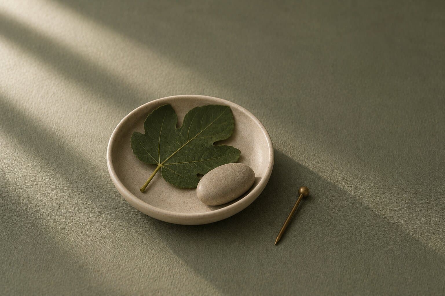 A green fig leaf and smooth stone in a small ceramic dish beside a brass marker pin on a muted sage surface